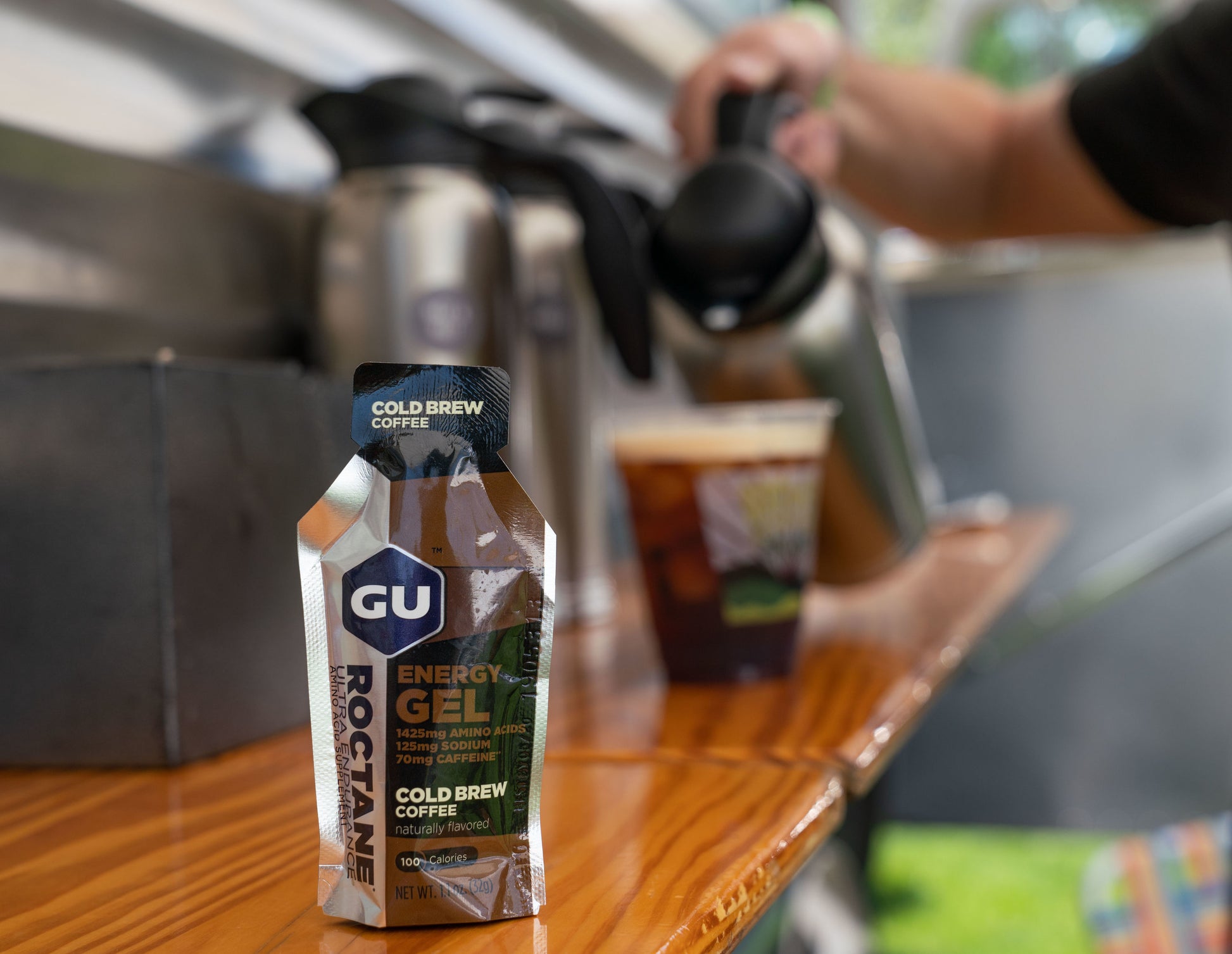Cold Brew Coffee Gu Energy gel in the forefront and a blurred background of person pouring milk into a coffee cup.