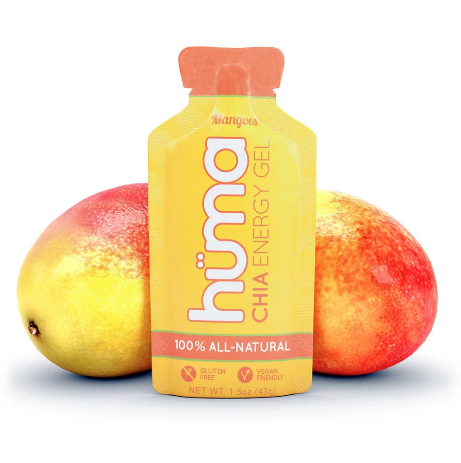 A packet of Huma Plus Gel with a mango flavor, alongside two whole mangoes. The gel packet is yellow with text and branding, indicating it is 100% all-natural.