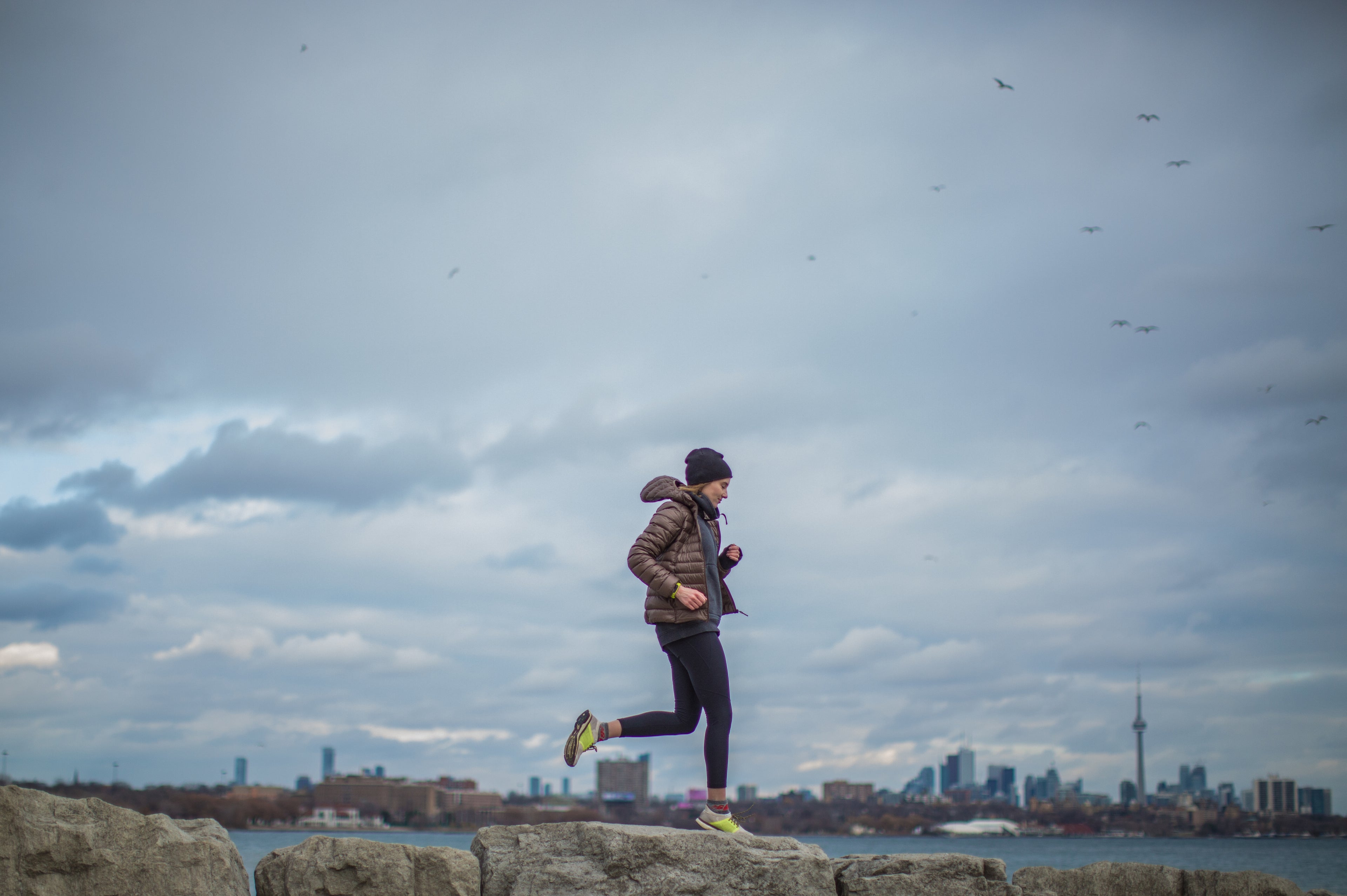 Runner in a jacket and hat jogging on rocks by the water with the Toronto skyline in the background under a cloudy sky