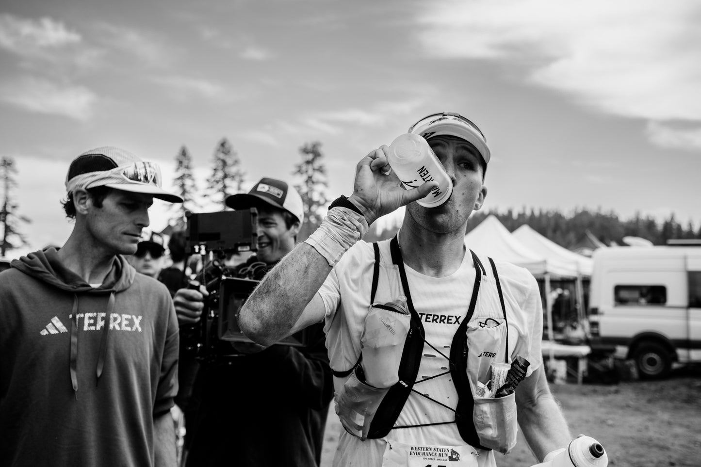 Male trail runner in a white hydration vest drinking from a clear Maurten water bottle with camper vans and people in background