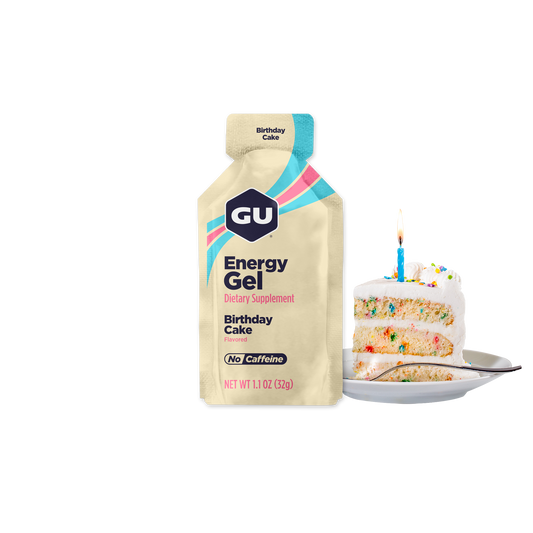 A packet of GU Energy Gel with Birthday Cake flavor, alongside a slice of birthday cake with a candle on it.