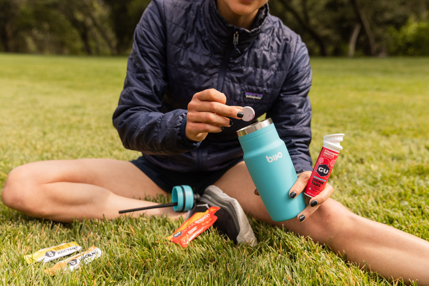 Runner sitting on grass in a coat putting a drink mix tablet in blue water bottle