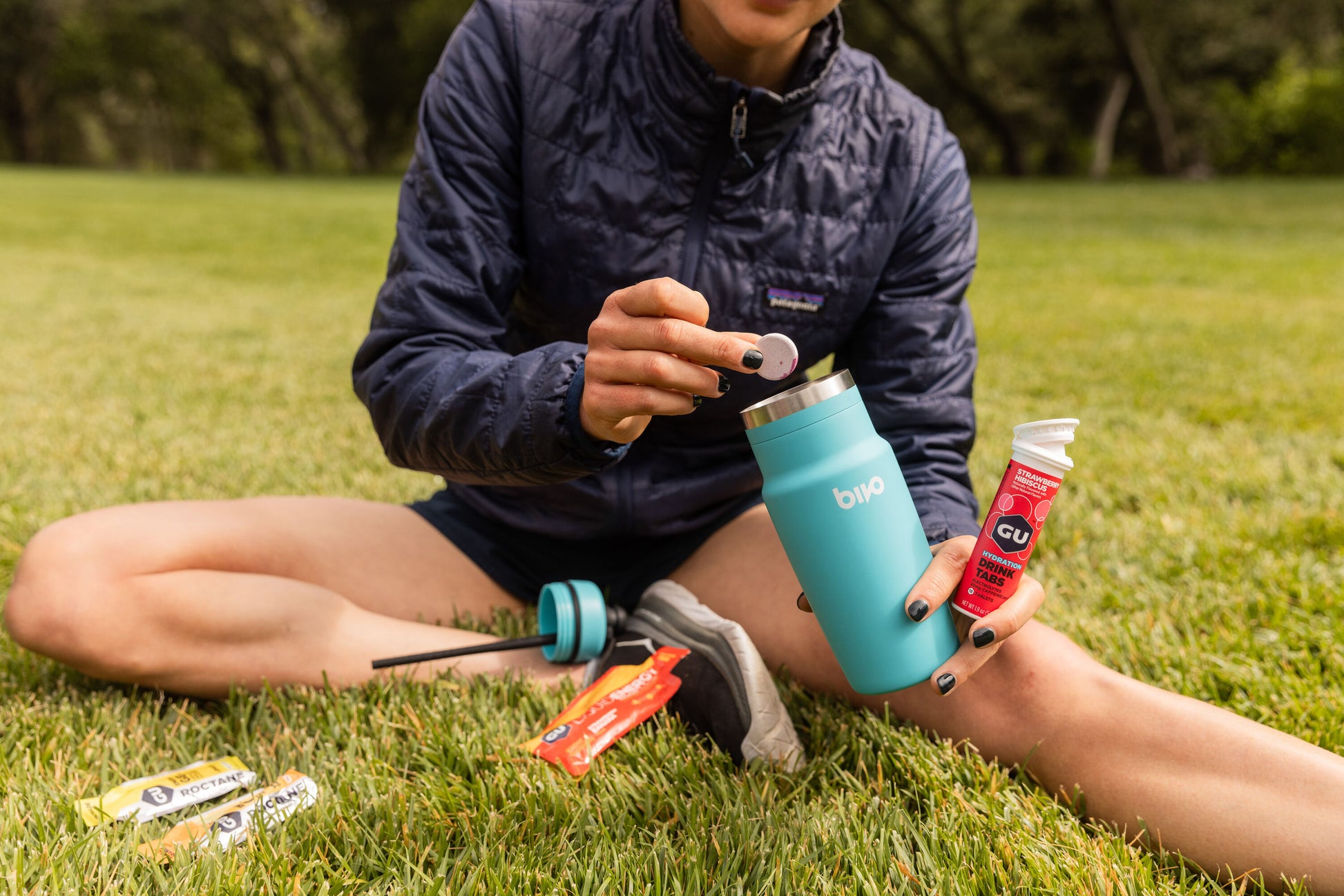 Runner sitting on grass in a coat putting a drink mix tablet in blue water bottle
