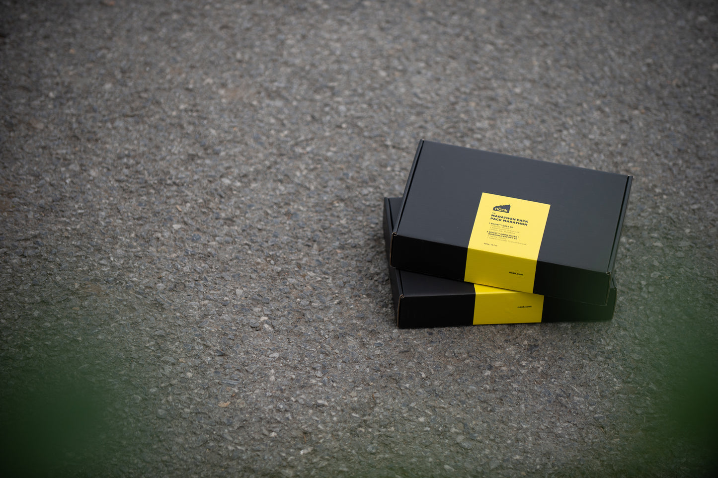 Two black rectangular naak Marathon Pack boxes stacked on a gray asphalt surface, each with a bright yellow label on the front | Fuel Kit