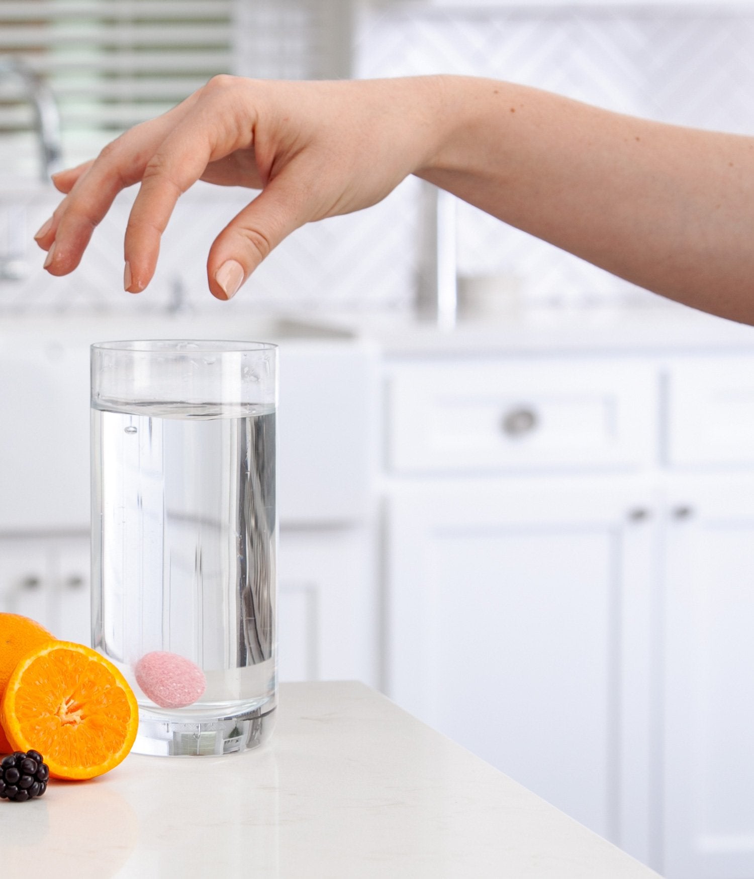 Glass of water with an orange, blackberry, and a pink tablet on a kitchen counter.