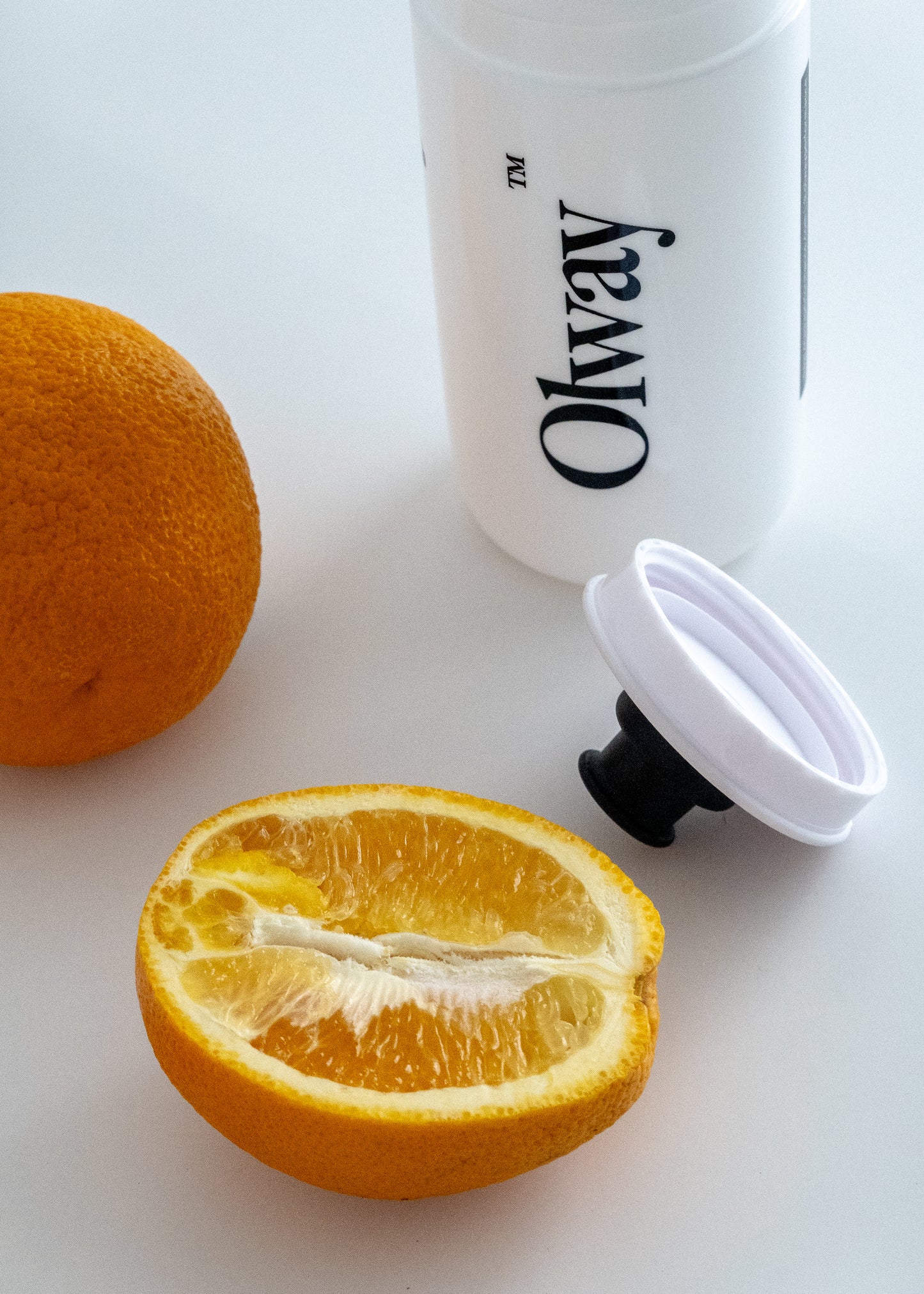 A white Olway water bottle with a black logo, shown with a whole orange, a halved orange, and a white cap available at Fuel Kit.