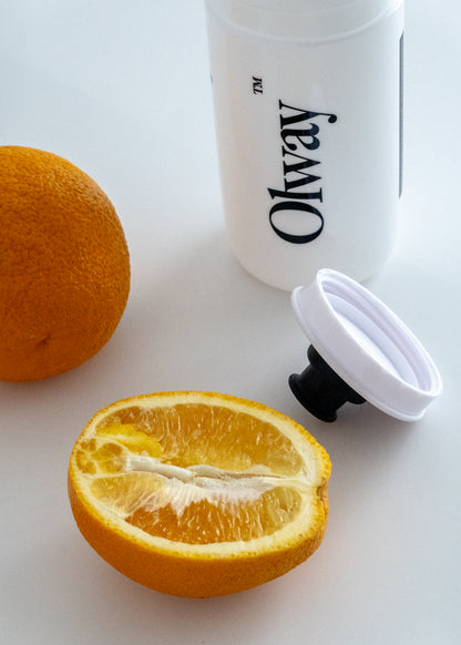 A white Olway water bottle with a black logo, shown with a whole orange, a halved orange, and a white cap available at Fuel Kit.