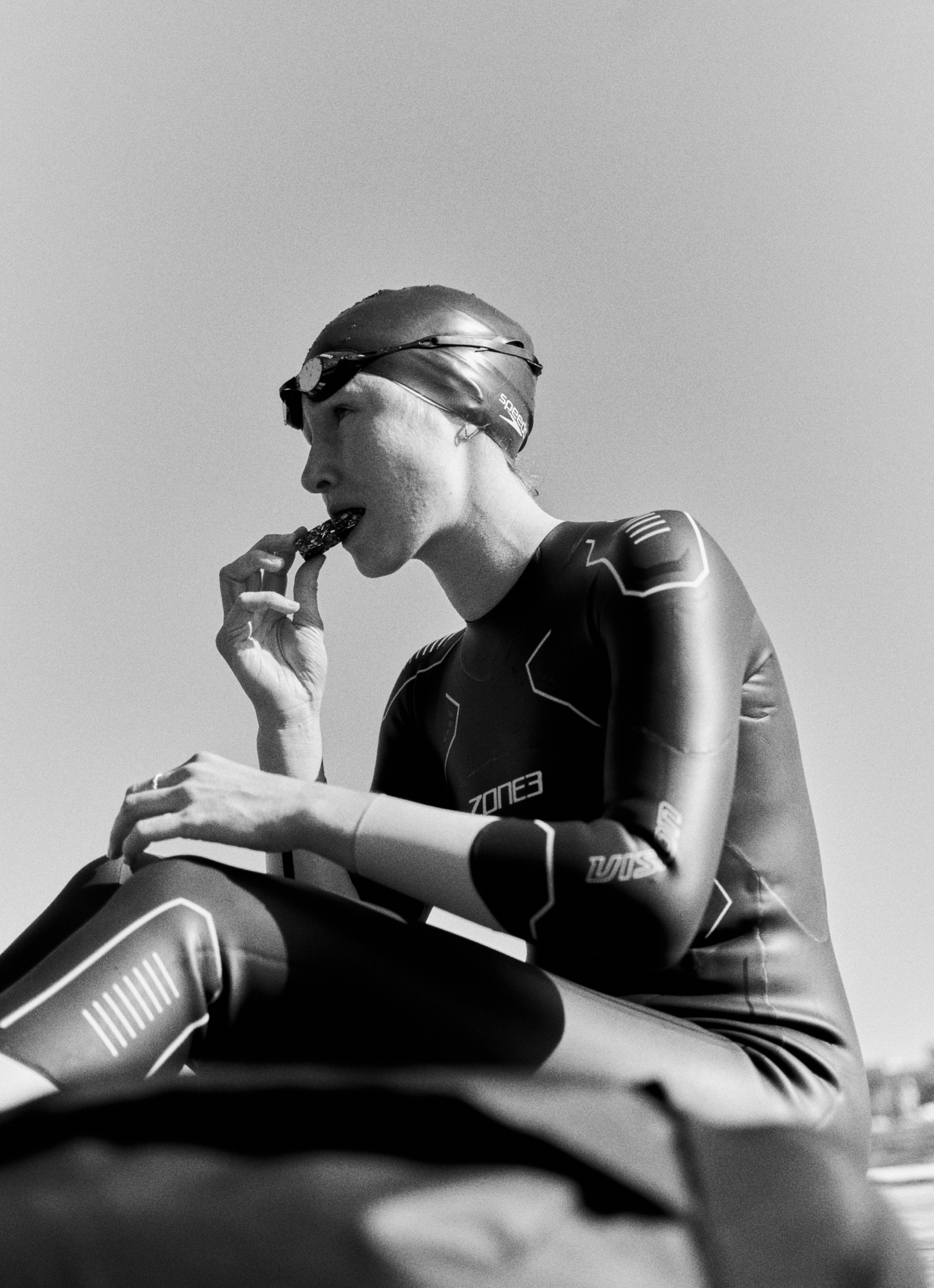 Triathlete wearing a wetsuit and swim cap sitting and eating a chocolate Maurten Solid Bar
