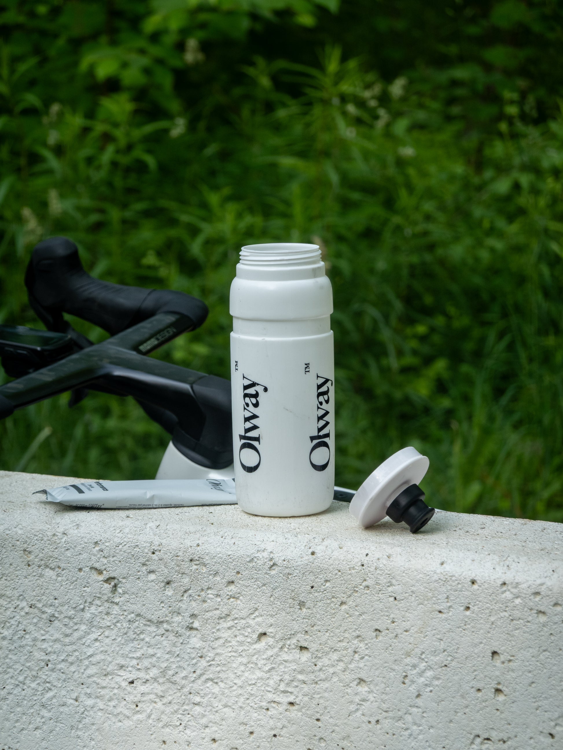 A white Olway cycling water bottle with a black bicycle handlebar visible behind it and green foliage in the background available at Fuel Kit